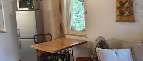 Un espace de vie compact comprenant une table à manger en bois, un canapé blanc, une kitchenette équipée d’un four à micro-ondes et une fenêtre donnant sur un paysage verdoyant.