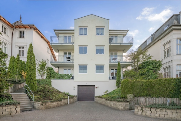 A multi-story residential building with a central entrance, balconies, and surrounding greenery.
