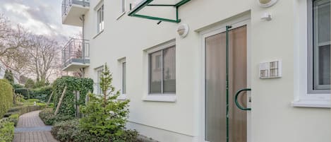 A modern building with a glass door entrance, a small garden area with a pathway, and a metal balcony.