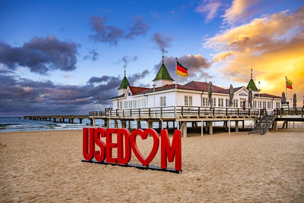 A beach with a large red sign reading 'USEDOM' and a building with multiple flags. - Cozy vacation apartment in Ahlbeck with sea view and balcony on Usedom (Ahlbeck)