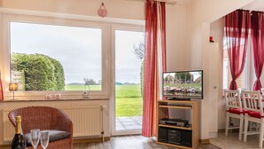 Ein gemütliches Wohnzimmer mit Fernseher, Korbsessel und Essbereich mit roten Kissen.