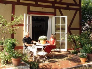 Un patio al aire libre con dos personas sentadas en una mesa, rodeado de macetas con plantas y un edificio con vigas de madera a la vista.