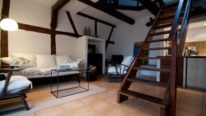 Ein gemütliches Wohnzimmer mit einer Couch, einem Couchtisch und einem Holzstuhl. Eine Holzleiter führt in einen Küchenbereich mit Kühlschrank und Kaffeemaschine.