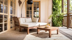 Eine Holzterrasse mit einer Couch und einem Tisch, ein großes Fenster mit Vorhängen und ein Blumentopf.