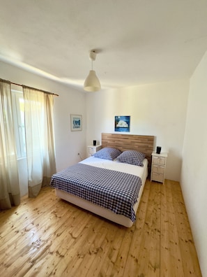 A bedroom with a wooden floor, a bed with checkered bedding, a white headboard, bedside tables with lamps, and a painting on the wall.