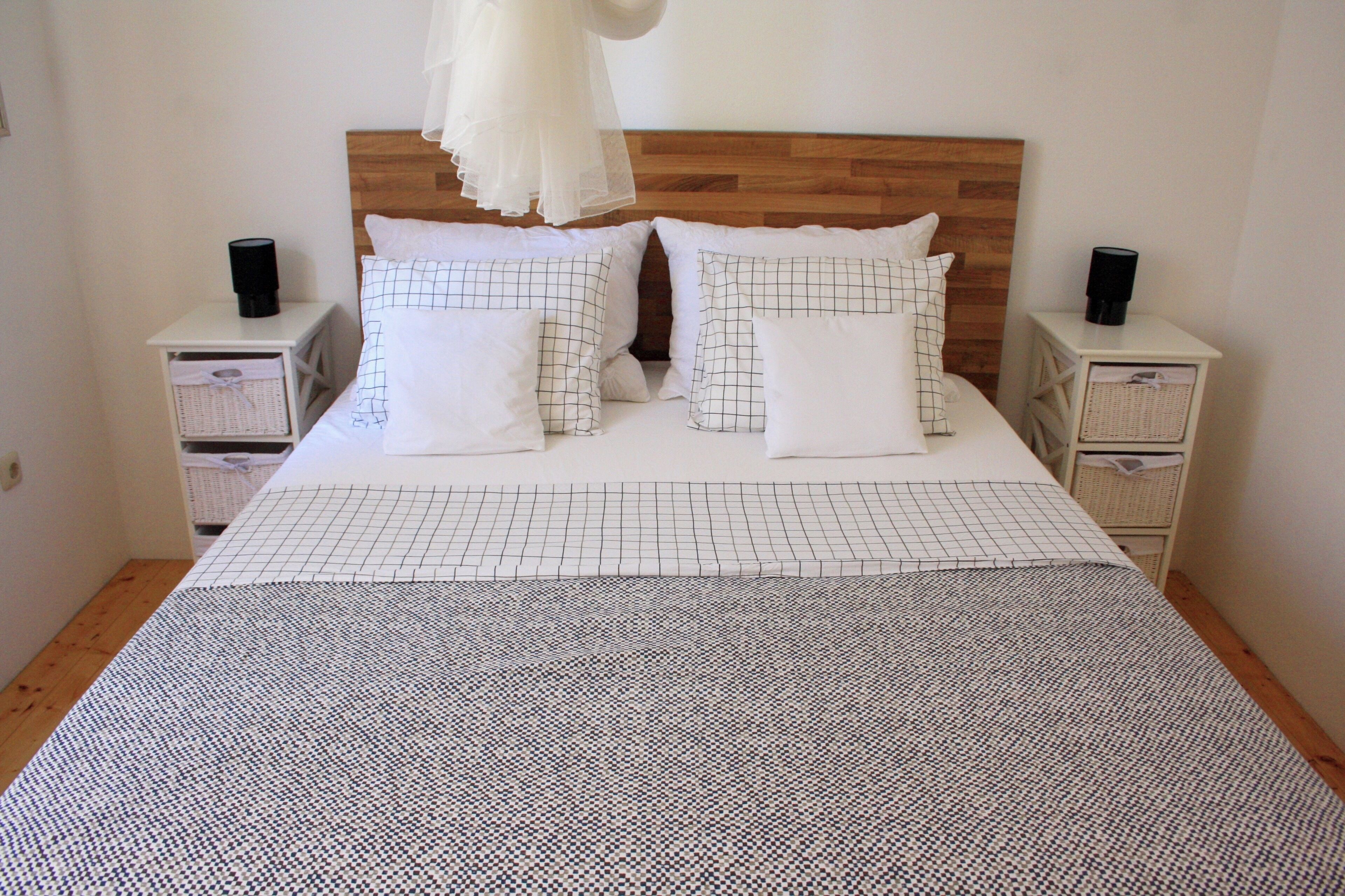 Una cama bien hecha con una colcha estampada, dos almohadas con diseño de cuadrícula y un cabecero de madera. Hay dos mesitas de noche con parlantes negros y cestas blancas.