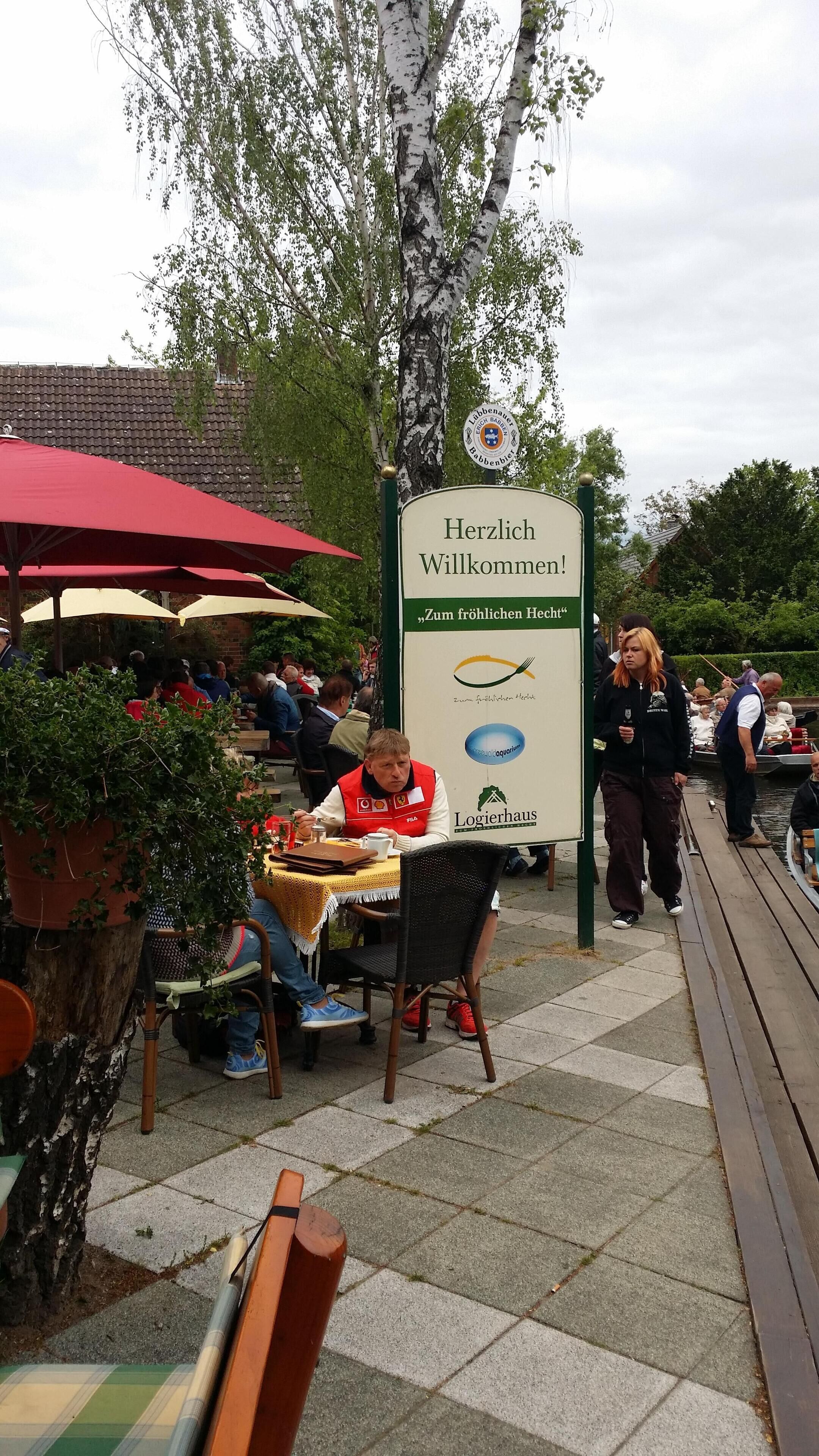 Ein Außenbereich zum Essen mit Tischen und Stühlen, ein Schild mit der Aufschrift 'Herzlich Willkommen!' und Blick auf einen Fluss mit Booten.