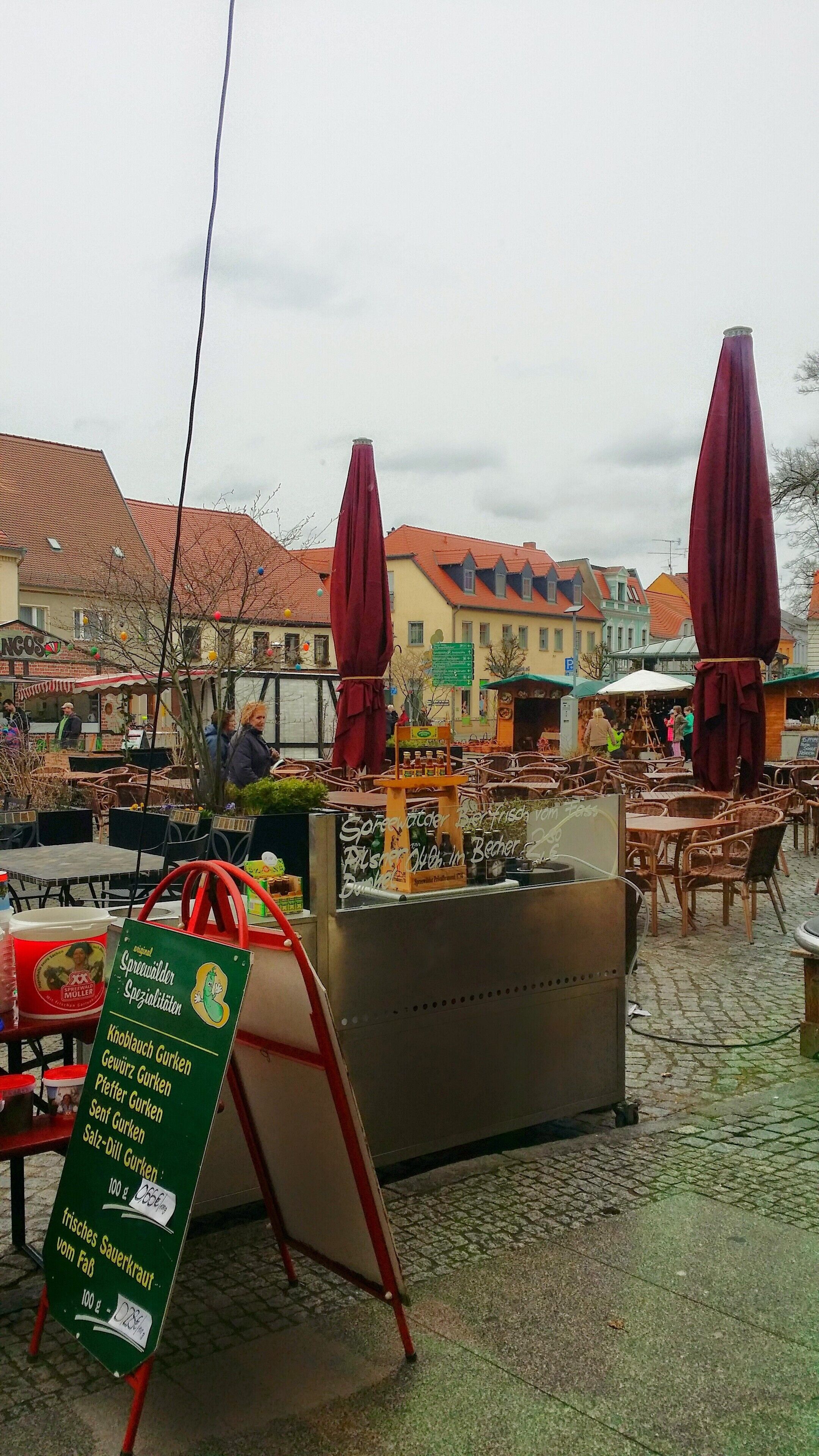 An outdoor café with tables and chairs, a signboard with menu items, and buildings in the background.