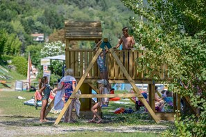 A wooden play structure with a slide and climbing area, surrounded by grass and trees.