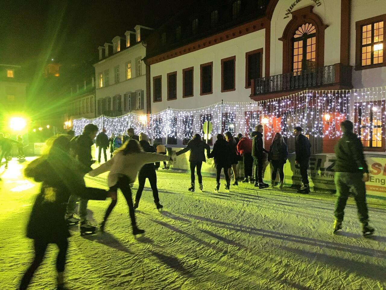 A group of people ice skating on a frozen surface in front of a building with string lights.