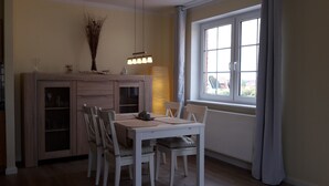 Un coin repas avec une table et des chaises blanches, un meuble en bois doté de portes en verre et une fenêtre avec des rideaux.