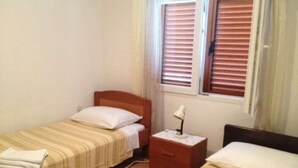 Une petite chambre propre, avec deux lits simples, une table de chevet avec une lampe et une fenêtre avec des stores.