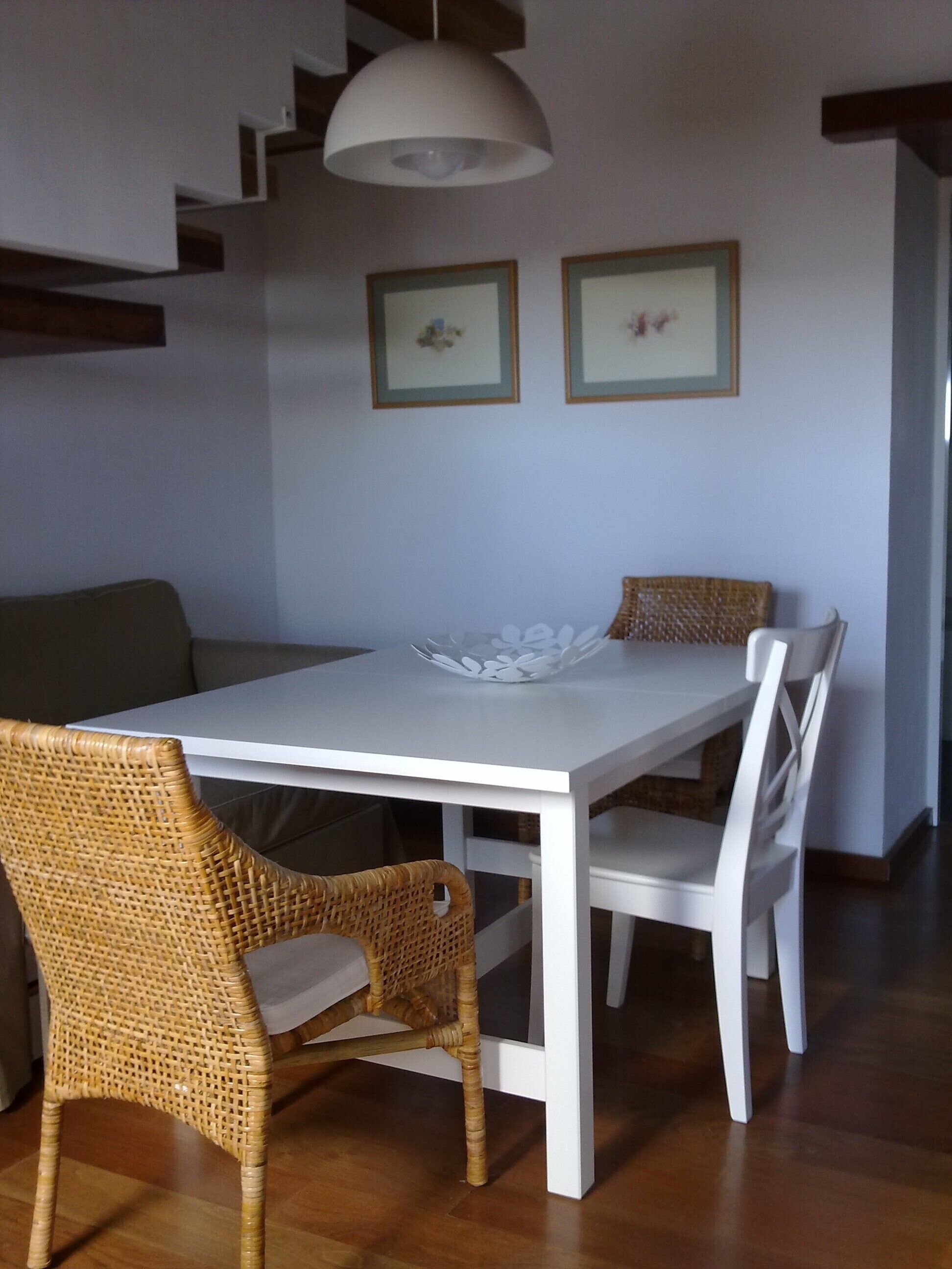 A dining area with a white table, wicker chairs, and a white bench. There are two framed pictures on the wall.
