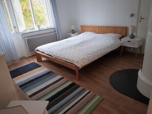 A bedroom with a wooden bed, a radiator, a window with curtains, a bedside table with a lamp, and a striped rug. - In the heart of Smaland hyggeliges cottage Klavreström with canoe! (Klavreström)