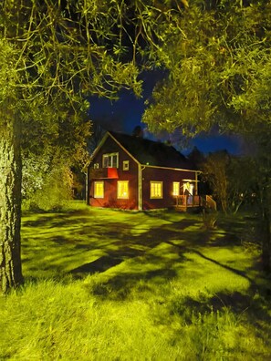 A house with a red exterior, a porch, and a well-lit front yard at night. - In the heart of Smaland hyggeliges cottage Klavreström with canoe! (Klavreström)