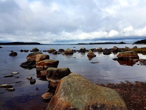 A rocky shoreline with calm water and distant trees under a cloudy sky. - In the heart of Smaland hyggeliges cottage Klavreström with canoe! (Klavreström)
