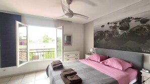 A bedroom with a bed, a ceiling fan, a window with a view of the outside, a nightstand, and a wall-mounted TV.