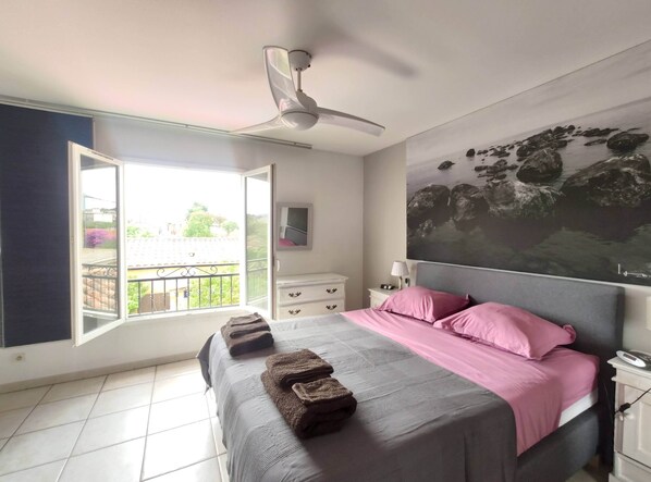 Une chambre à coucher comprenant un lit, un ventilateur de plafond, une fenêtre donnant sur l’extérieur, une table de chevet et un téléviseur fixé au mur.