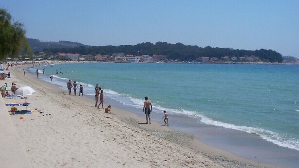 A beach with people walking along the shore, umbrellas, and a clear view of the sea and distant hills.