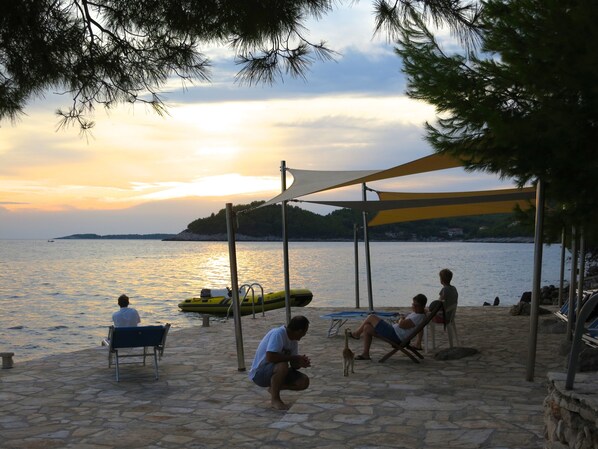 A beachside setting with a stone-paved area, a yellow kayak, and people relaxing under a shaded area. - Top apartment for 4-5, south. Location, right on the sea, boat dock, bike hit! (Blato)