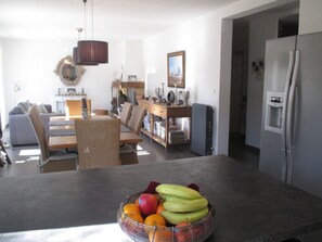 A modern kitchen with a fruit bowl on the counter, a dining area with a sofa and chairs, and a refrigerator.