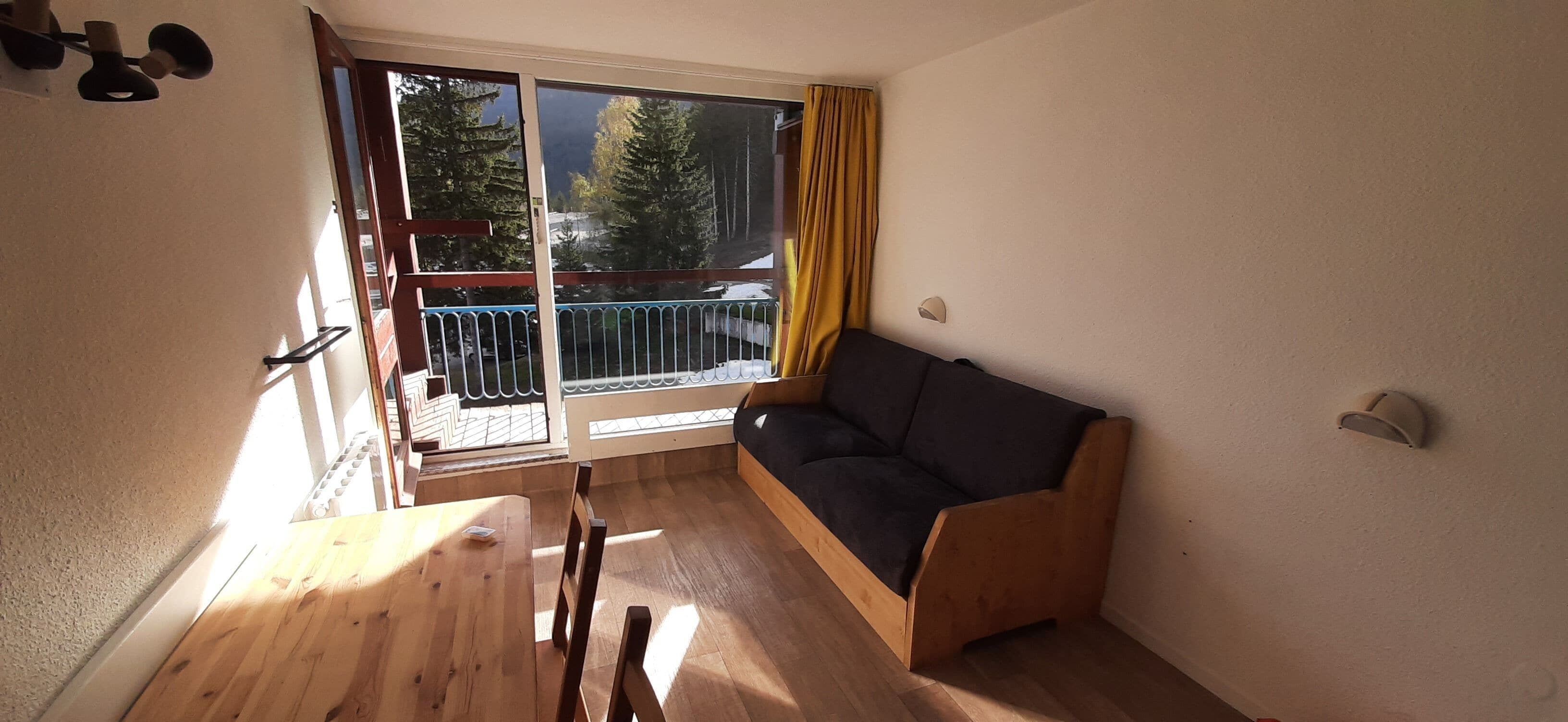 A room with a wooden table, a sofa, a sliding glass door leading to a balcony, and a view of trees.