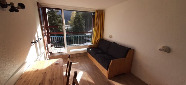 A room with a wooden table, a sofa, a sliding glass door leading to a balcony, and a view of trees.