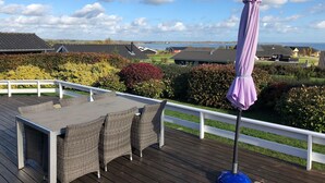 En træterrasse med udendørs møbler, en lilla parasol og udsigt til huse og vand i baggrunden.