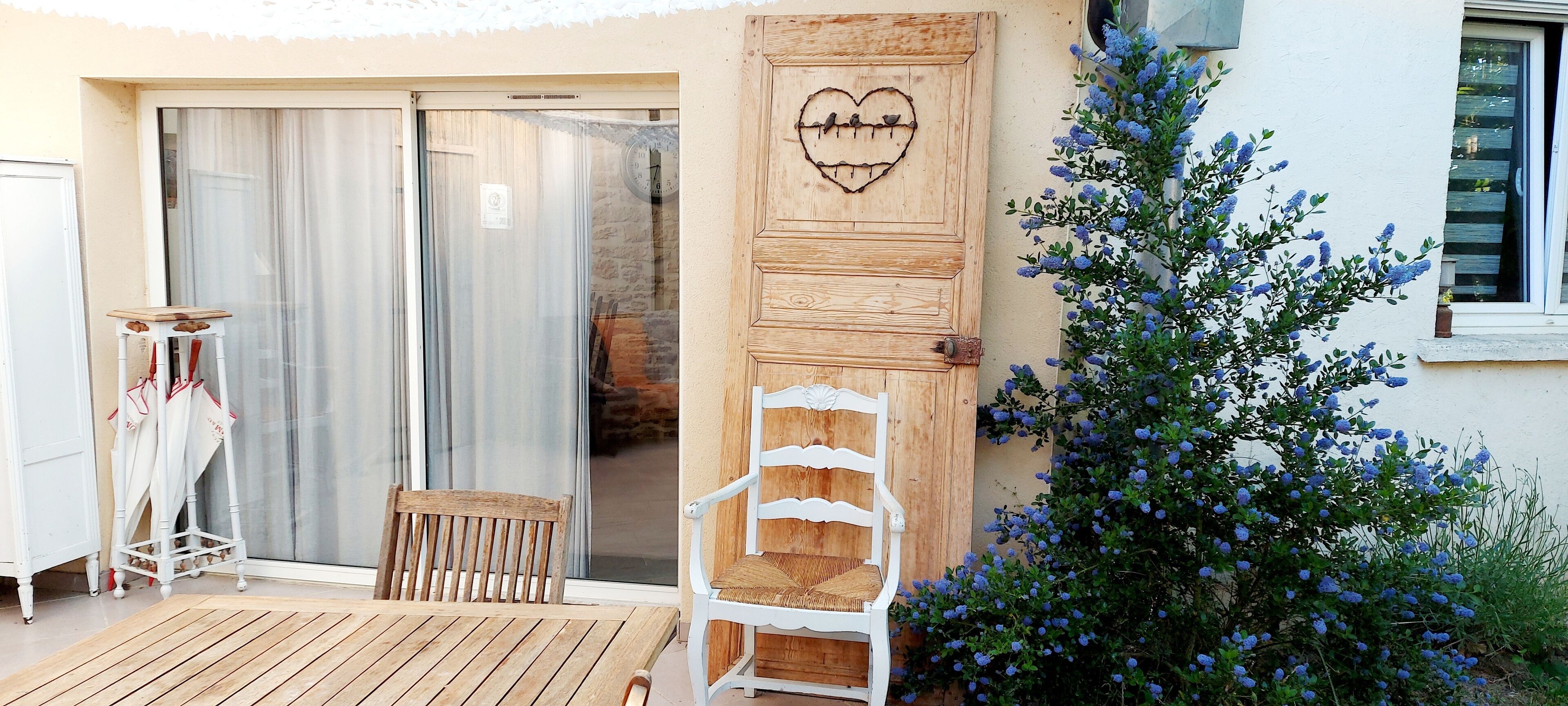 A wooden deck with a white chair and a small table, a sliding glass door with a heart-shaped decoration, and a bush with blue flowers.