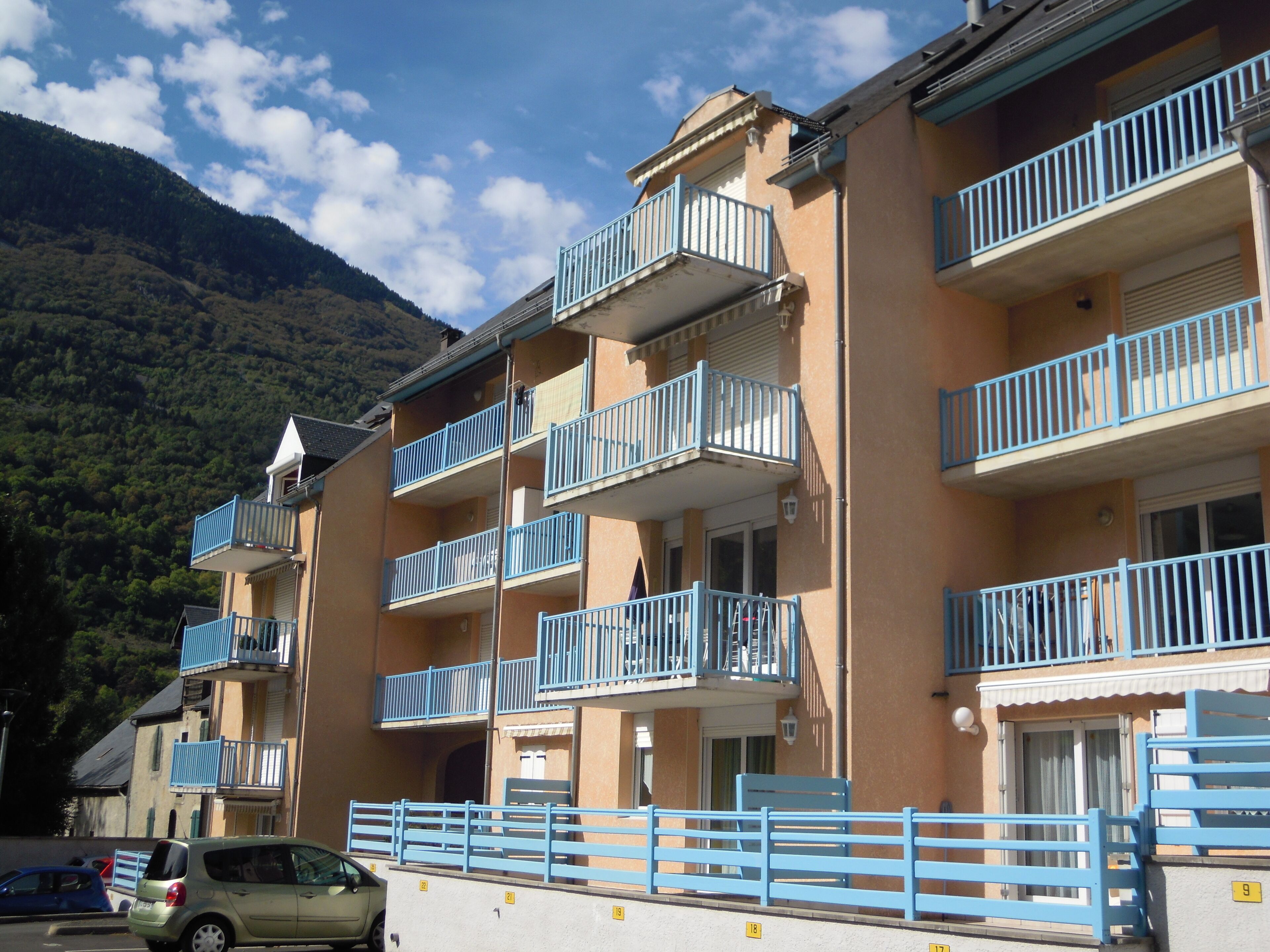 A multi-story building with balconies, a car parked in front, and a mountainous backdrop.