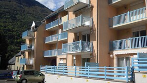 A multi-story building with balconies, a car parked in front, and a mountainous backdrop.