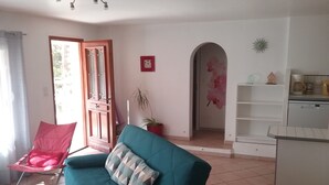 Un salon avec un canapé de couleur turquoise, un fauteuil rouge, une plante en pot, un coin cuisine doté d’un plan de travail blanc, et une porte donnant sur une autre pièce.