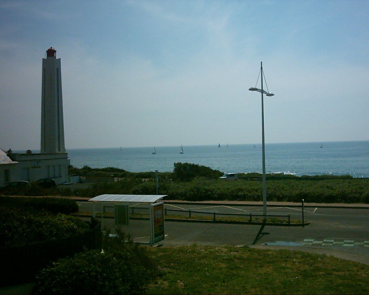 A coastal view with a lighthouse, a bus stop, and sailboats on the water.