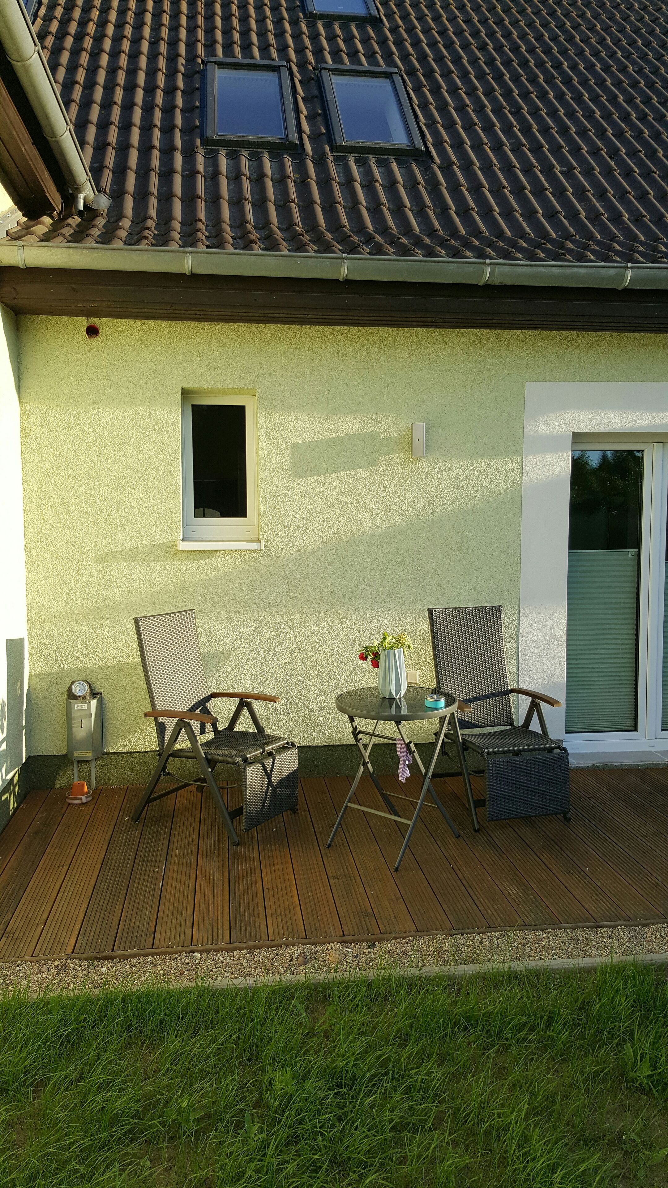 Eine überdachte Terrasse mit zwei Korbstühlen, einem kleinen Tisch mit einer Vase und einem Fenster.