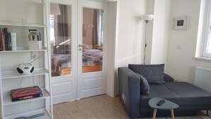 A room with a grey sofa, a small table, a bookshelf, and a door leading to a bedroom. - New apartment in the middle of the lake district ideal for active recreation (Blankensee)
