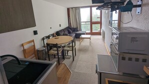 A compact living space with a kitchenette, a dining area with a round table and chairs, a sofa, and a large window offering a view of the outdoors.