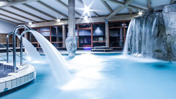 An indoor swimming pool with multiple water features, including a waterfall and a central fountain.