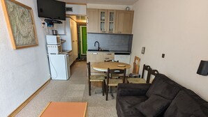 A compact living space with a black sofa, a wooden coffee table, a dining table with chairs, a kitchenette, and a television mounted on the wall.