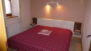 A bedroom with a red checkered bedspread, a nightstand with a lamp, and a window with curtains. - highland cottage (Grendelbruch)