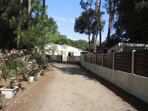 A pathway leading to a white building with a fenced area, surrounded by trees and flowering plants. - Villa, Jard sur mer (Vendée), 3 Bedrooms, 110m² (1,184 sq ft) (Jard-Sur-Mer)