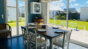 A dining area with a table set for four, white chairs, a wooden table, and a view of a green lawn and houses through large glass doors.