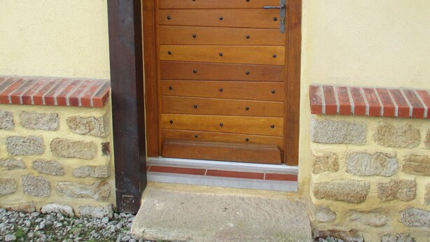 Une porte en bois, dotée d’une poignée en métal, est encastrée dans un mur en pierre et en briques, et un petit marchepied mène à son seuil.
