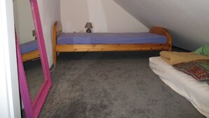 Une petite chambre avec un lit simple, un plafond en pente, un cadre de lit en bois, une petite table de chevet avec une lampe et un miroir sur le côté gauche.