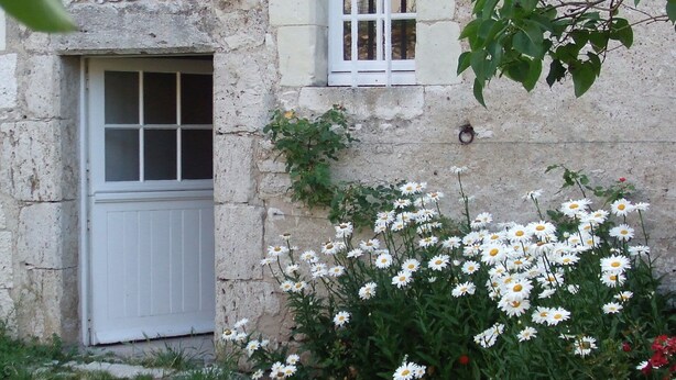 Edificio de piedra con puerta y ventana blanca, rodeado de follaje y flores blancas.