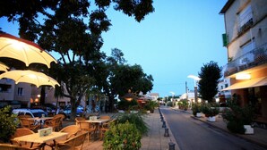 An outdoor cafe with wicker tables and chairs, umbrellas, and potted plants along a street. - Welcome to U MERULU of "Vacances-a-moriani", a charming corsican apartment for 6 (San-Nicolao)