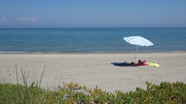 A beach with a person lying on a towel under a striped umbrella. - Welcome to U MERULU of "Vacances-a-moriani", a charming corsican apartment for 6 (San-Nicolao)