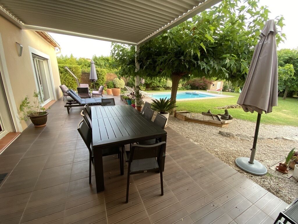 Un patio exterior con mesa y sillas de madera, rodeado de plantas en macetas y una piscina.
