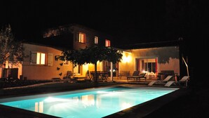 A nighttime view of a house with a swimming pool, outdoor seating, and warm lighting.