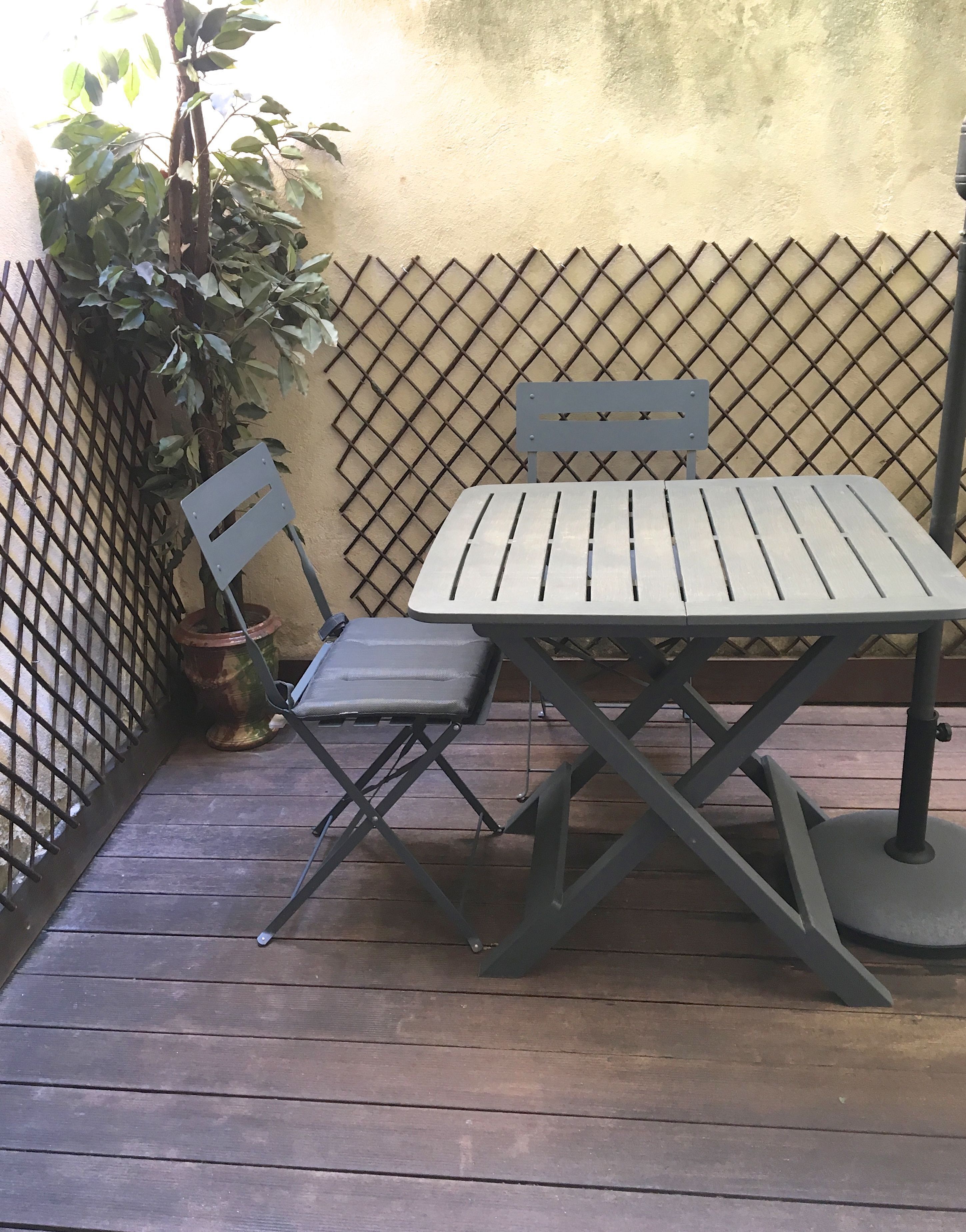 A small outdoor patio with a metal table and two chairs, a potted plant, and a metal fence.