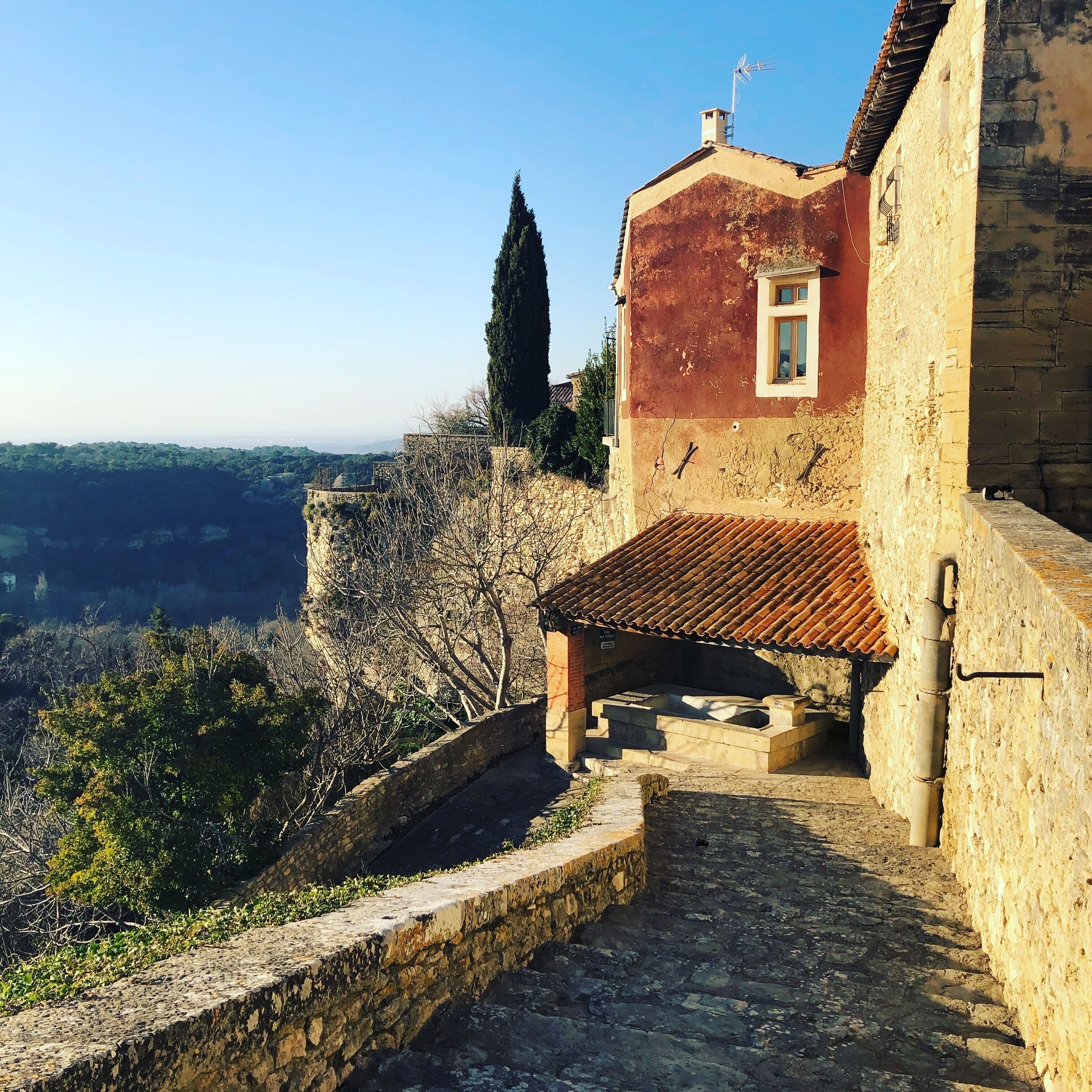 Ein Steinhaus mit rotem Ziegeldach und einem kleinen Fenster. Eine Steintreppe führt zu einer Plattform mit einer Steinsitzbank.